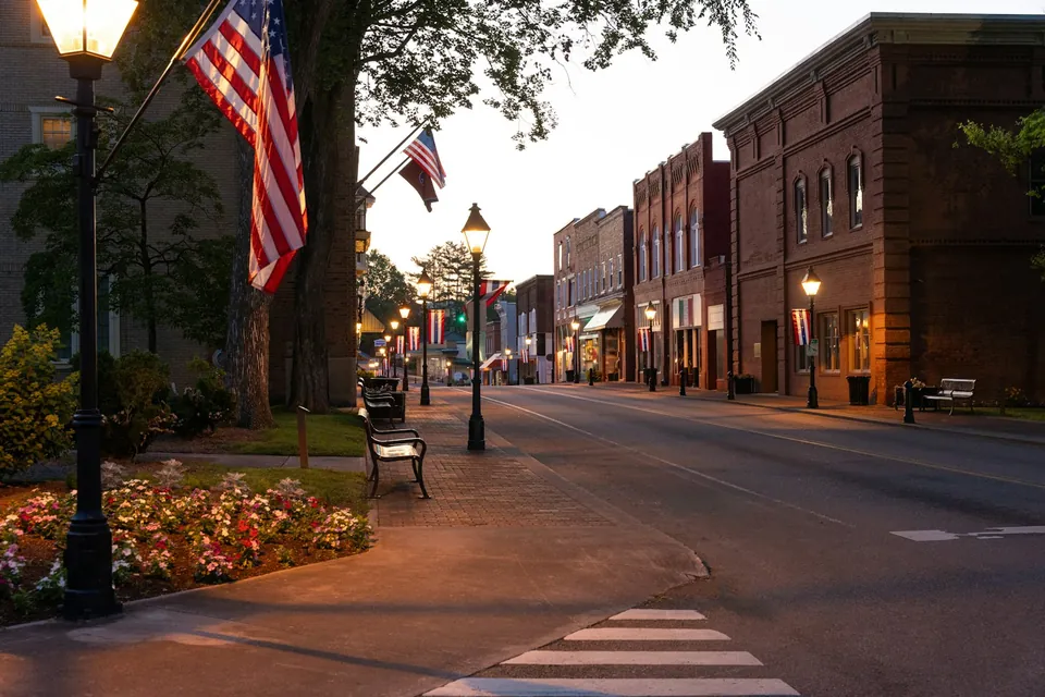 Brick storefronts, lamp posts and American flags along a small-town main street at dusk