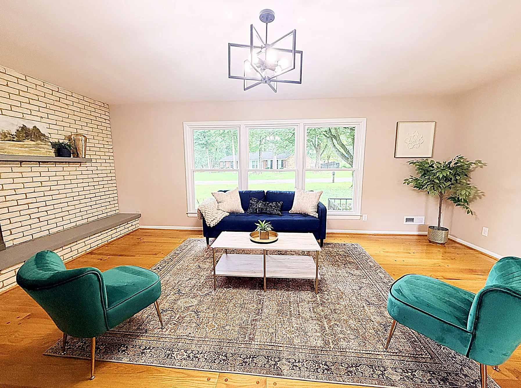 Living room with painted-white brick fireplace, navy sofa, emerald chairs, and a wide picture window over real hardwood floors