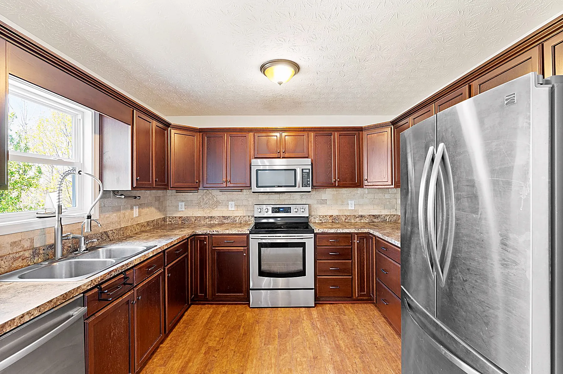 Wide view of the kitchen with a center island, granite counters, tile backsplash, and stainless steel appliances open to the living and dining space