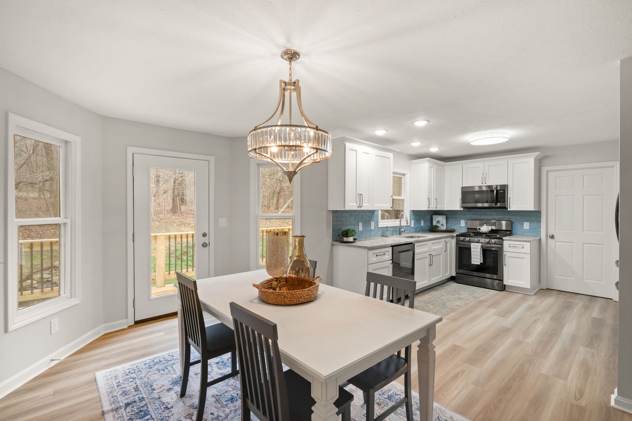 Open-concept kitchen and dining area with crystal chandelier, blue subway tile backsplash, and wooded deck views